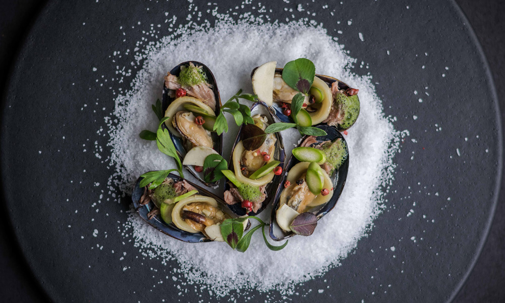 Black clam shells filled with mussels, herbs, microgreens and red roe, arranged on coarse salt on a dark plate.