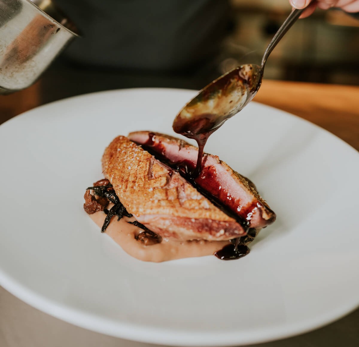 Sliced seared duck breast with crispy skin on a puree and mushrooms, chef spooning a glossy dark sauce over it on a white plate.