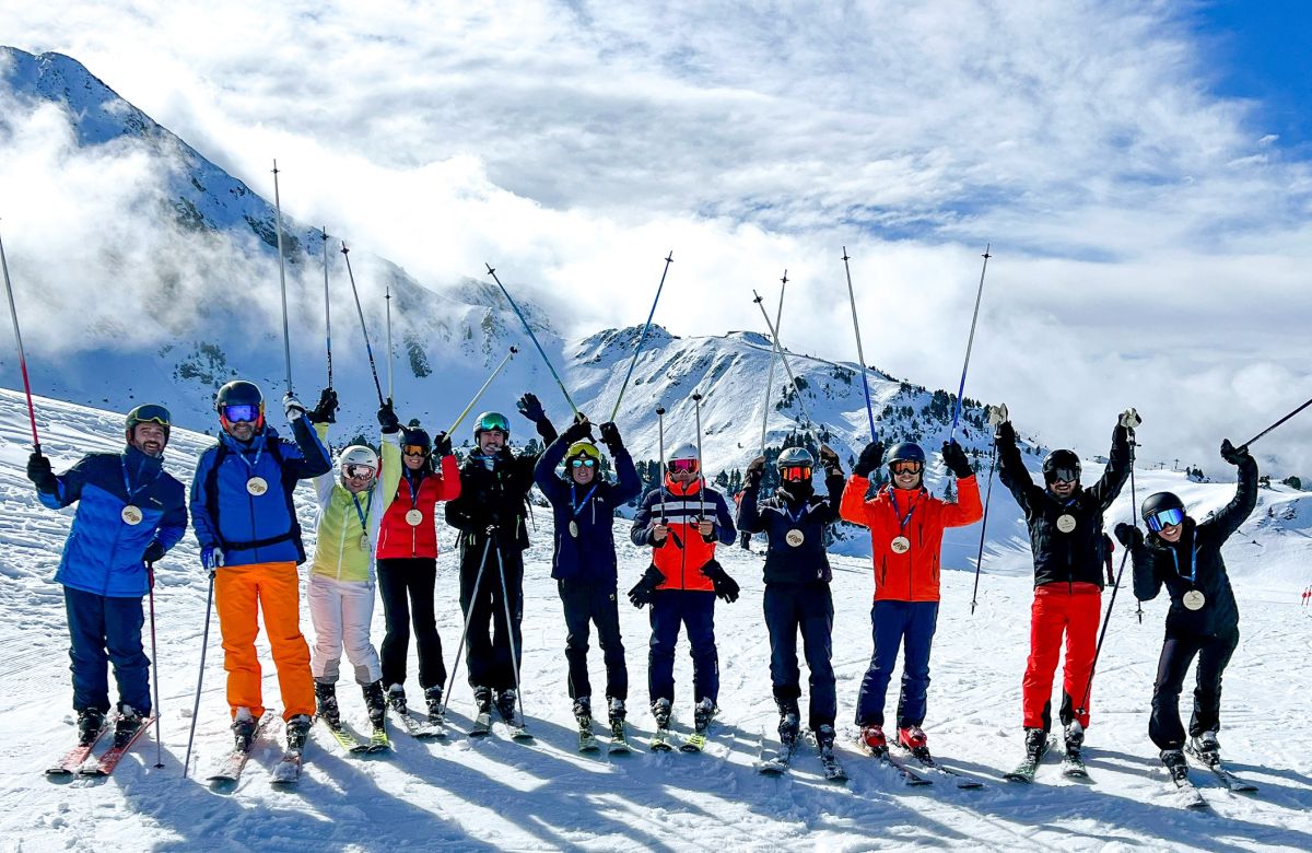 Group of skiers in colorful outfits pose joyfully on a snowy mountain slope, with ski poles raised against a backdrop of rugged peaks and cloudy sky.