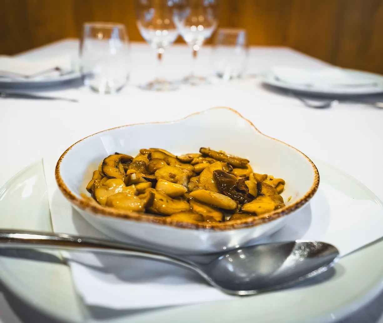 Sautéed mushrooms in a white bowl on a plate with a spoon, placed on a white tablecloth with blurred wine glasses behind.
