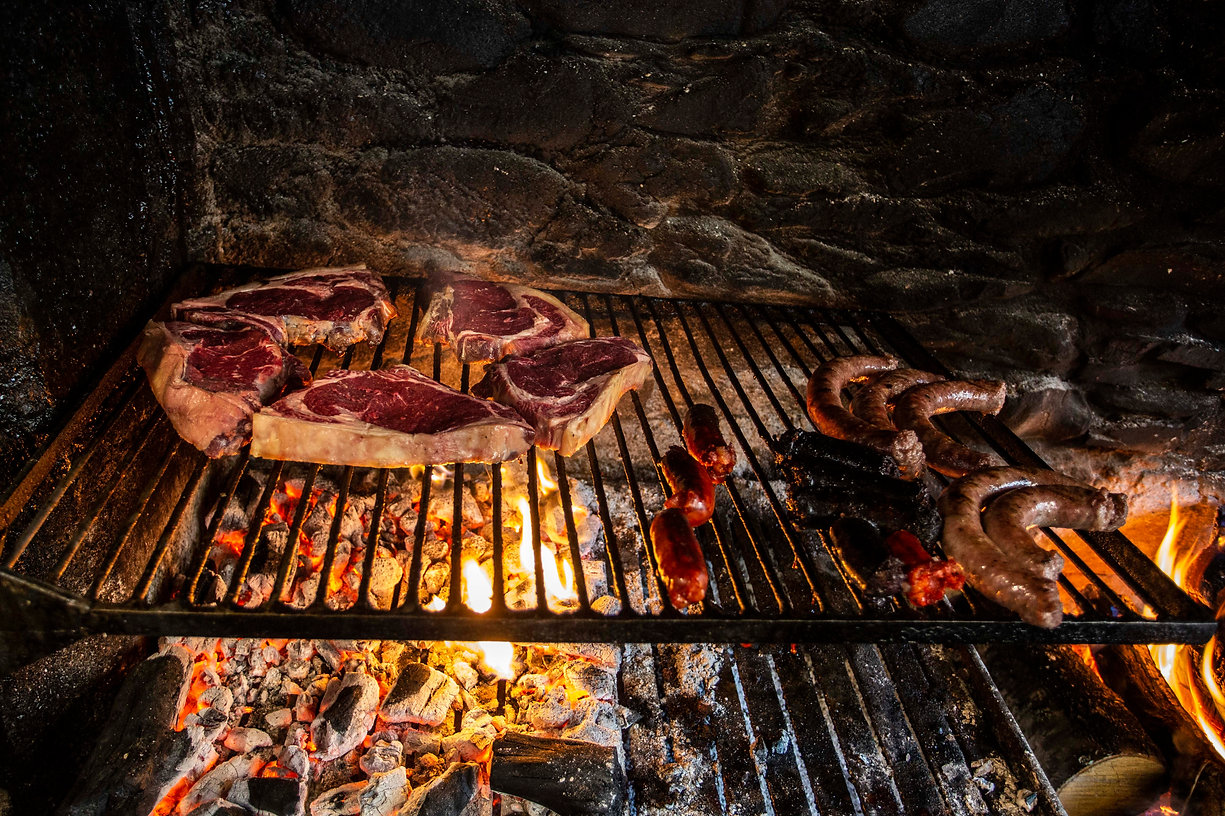 Thick ribeye steaks and sausages sizzling on a metal grill over glowing coals and flames in a stone pit.