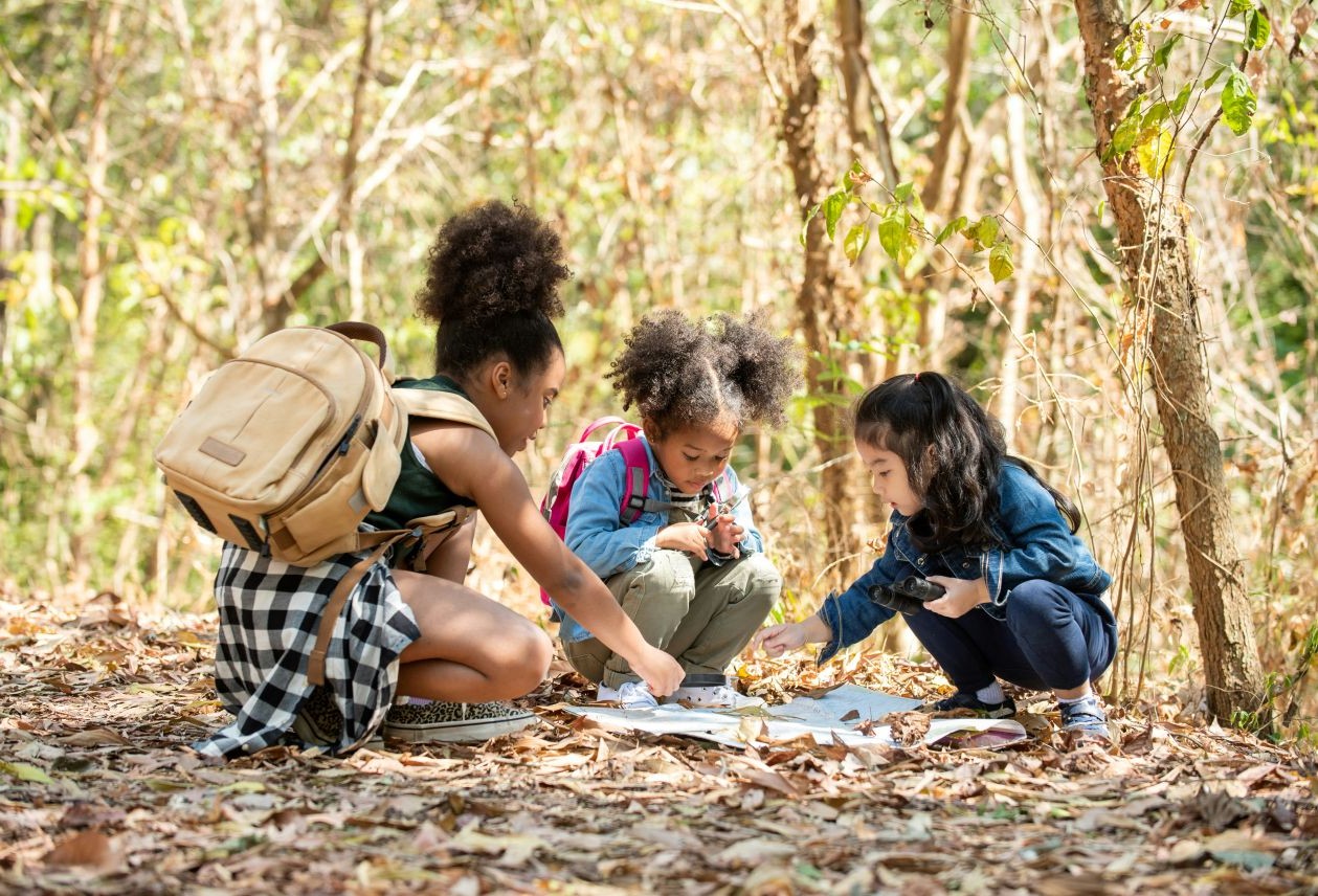 Three children crouched on a leafy forest floor, examining a map and nature items, backpacks nearby.