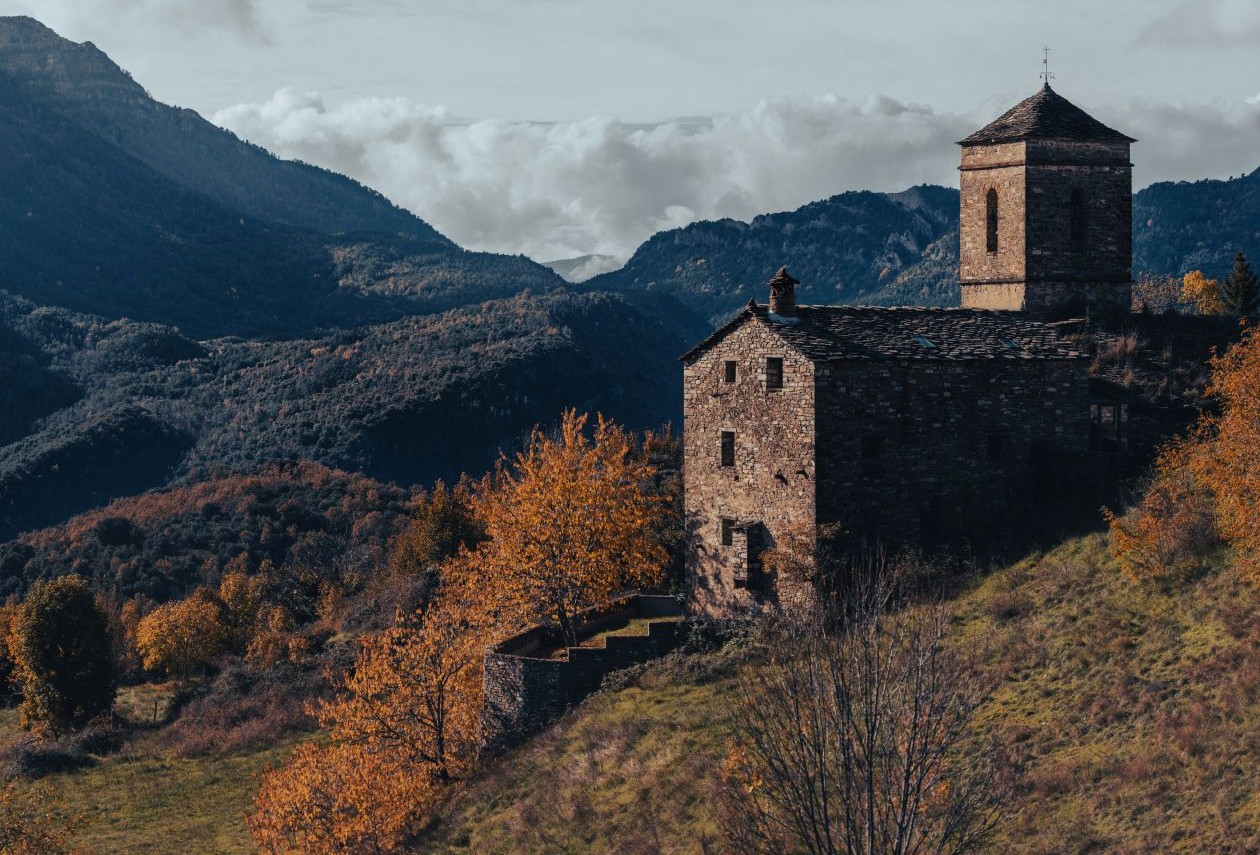 Old stone chapel with a tower on a grassy hillside, surrounded by autumn trees and layered mountains beneath a cloudy sky.