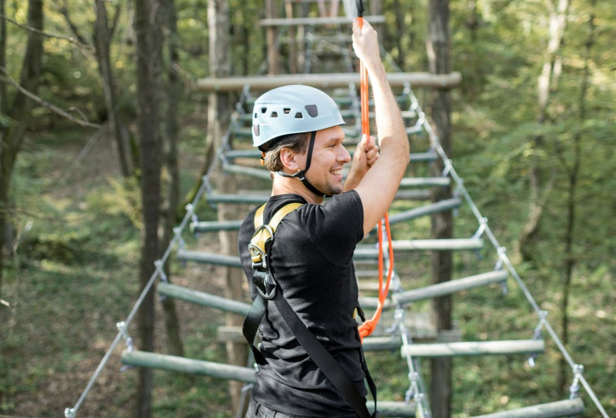 Smiling man in helmet and safety harness gripping an orange rope on a treetop rope-course ladder in a forest.