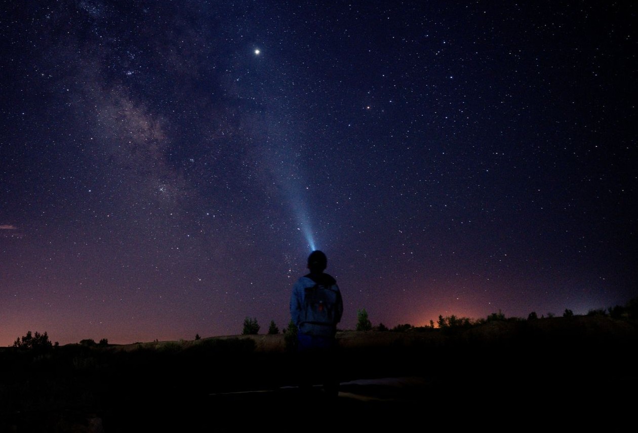 Silhouetted figure shining a headlamp beam into a star-filled sky with the Milky Way visible.