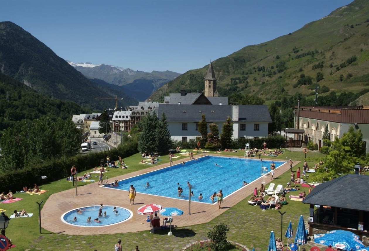 Outdoor hotel pool complex with swimmers and sunbathers on a grassy terrace, mountain valley and church steeple in the background.