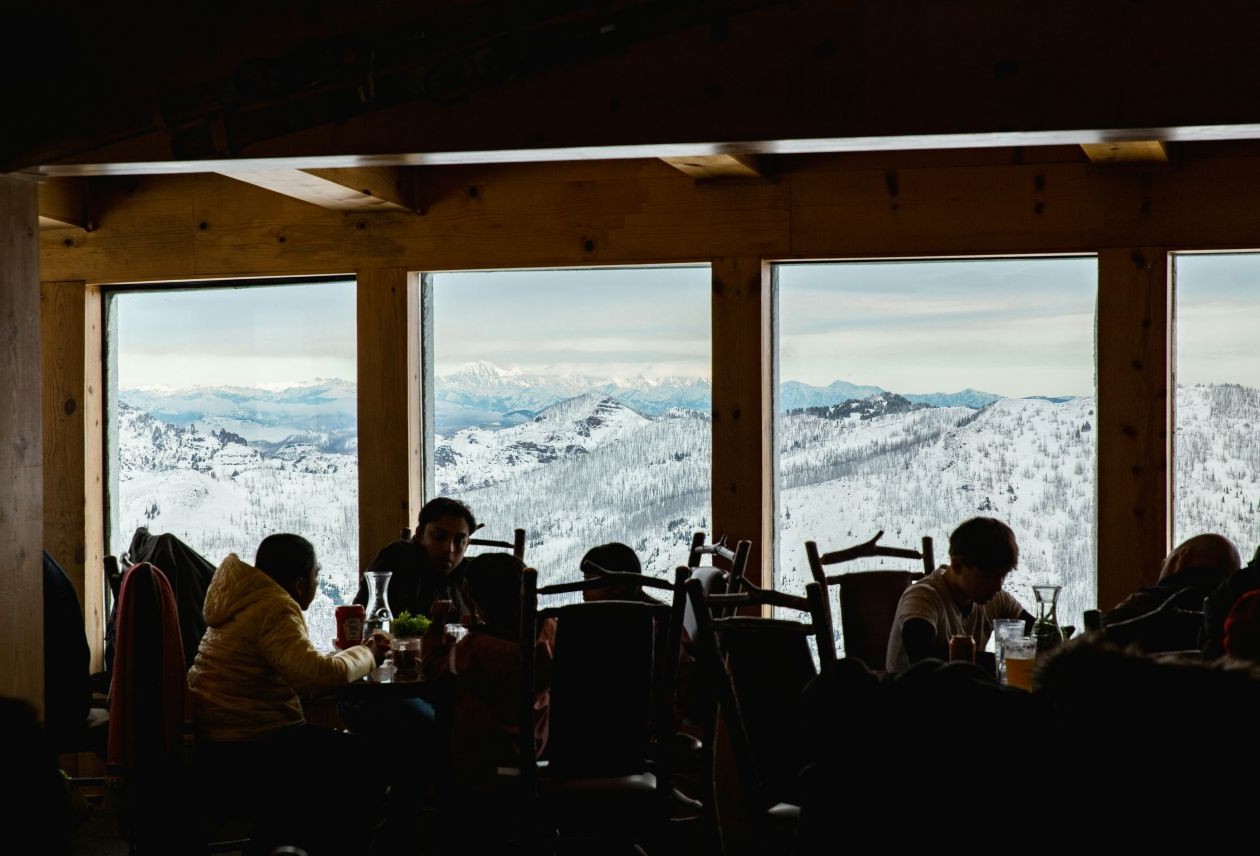 People dining in a wooden mountain lodge, silhouetted against large windows framing snow-covered peaks and valleys.