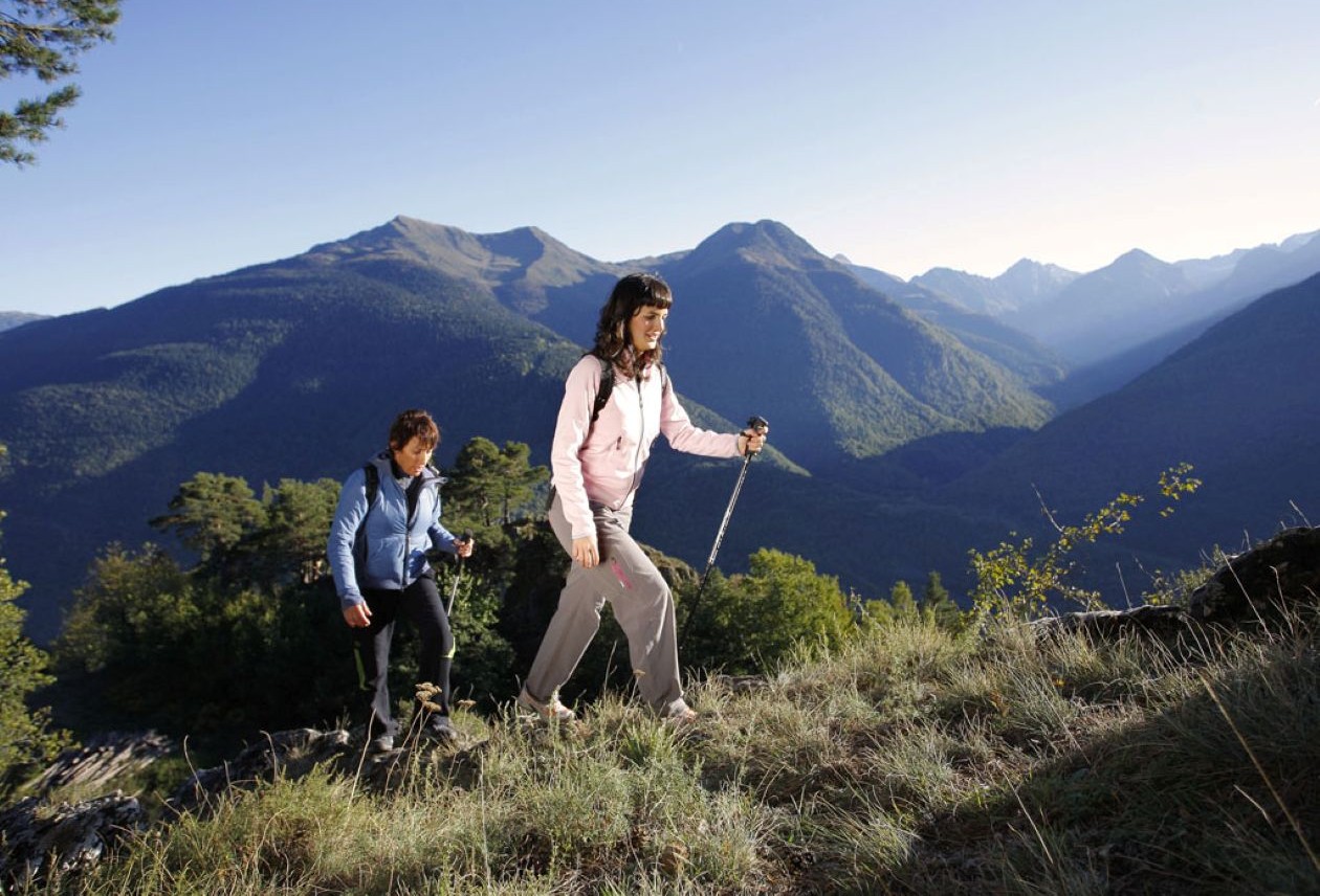 Two women hikers using trekking poles ascend a grassy ridge with forested mountains and valleys under a clear blue sky.