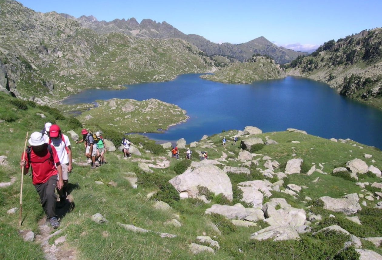 Group of hikers climbing a rocky, grassy slope toward a blue mountain lake and surrounding peaks.