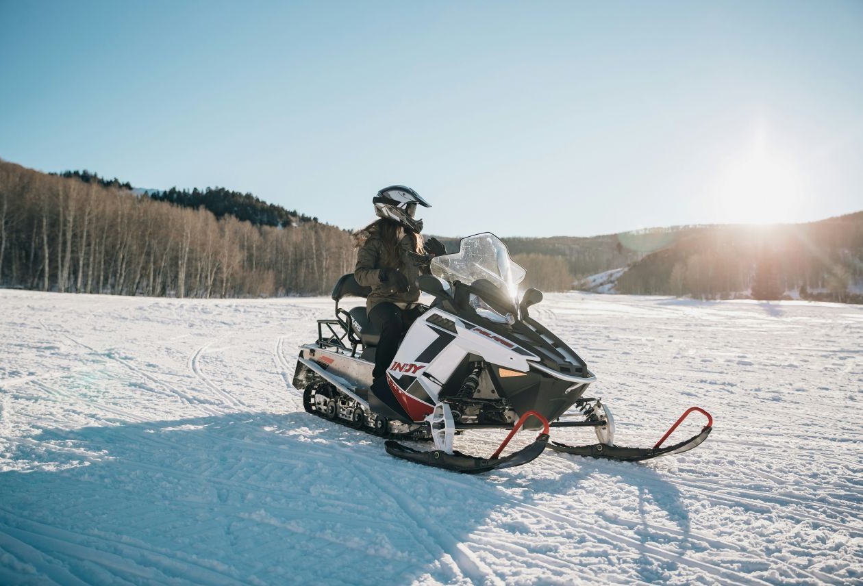Person in helmet seated on white-and-red snowmobile on a snowy plain, low sun and forested hills in the background.