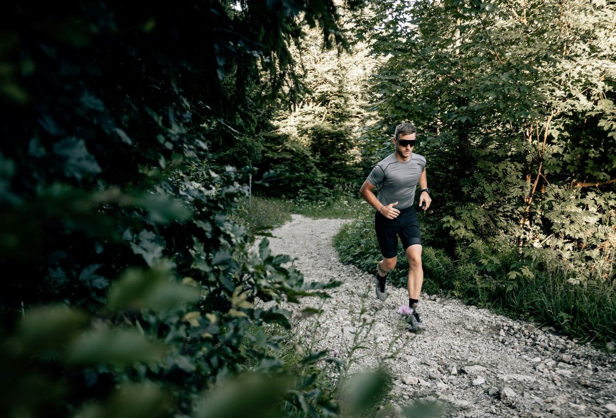 Man in sunglasses and athletic gear running on a rocky forest trail surrounded by dense green trees.