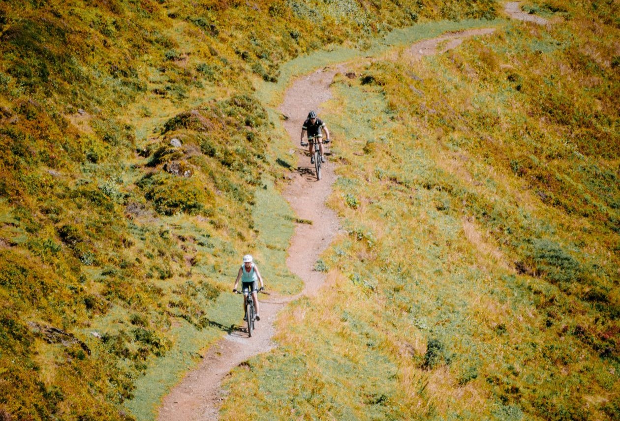 Two cyclists ride a narrow dirt trail winding through sunlit grassy hills, seen from above.