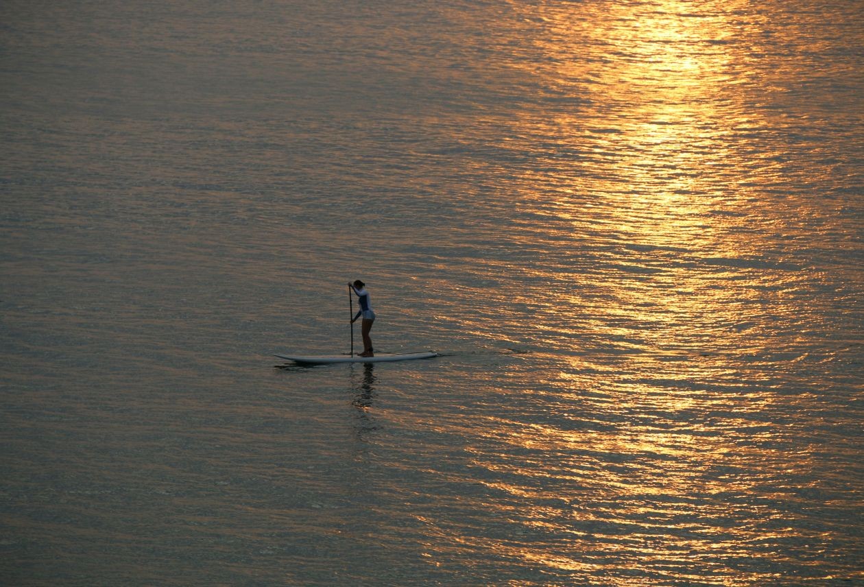 Solo paddleboarder gliding across calm water, silhouetted against a golden sunset reflection.