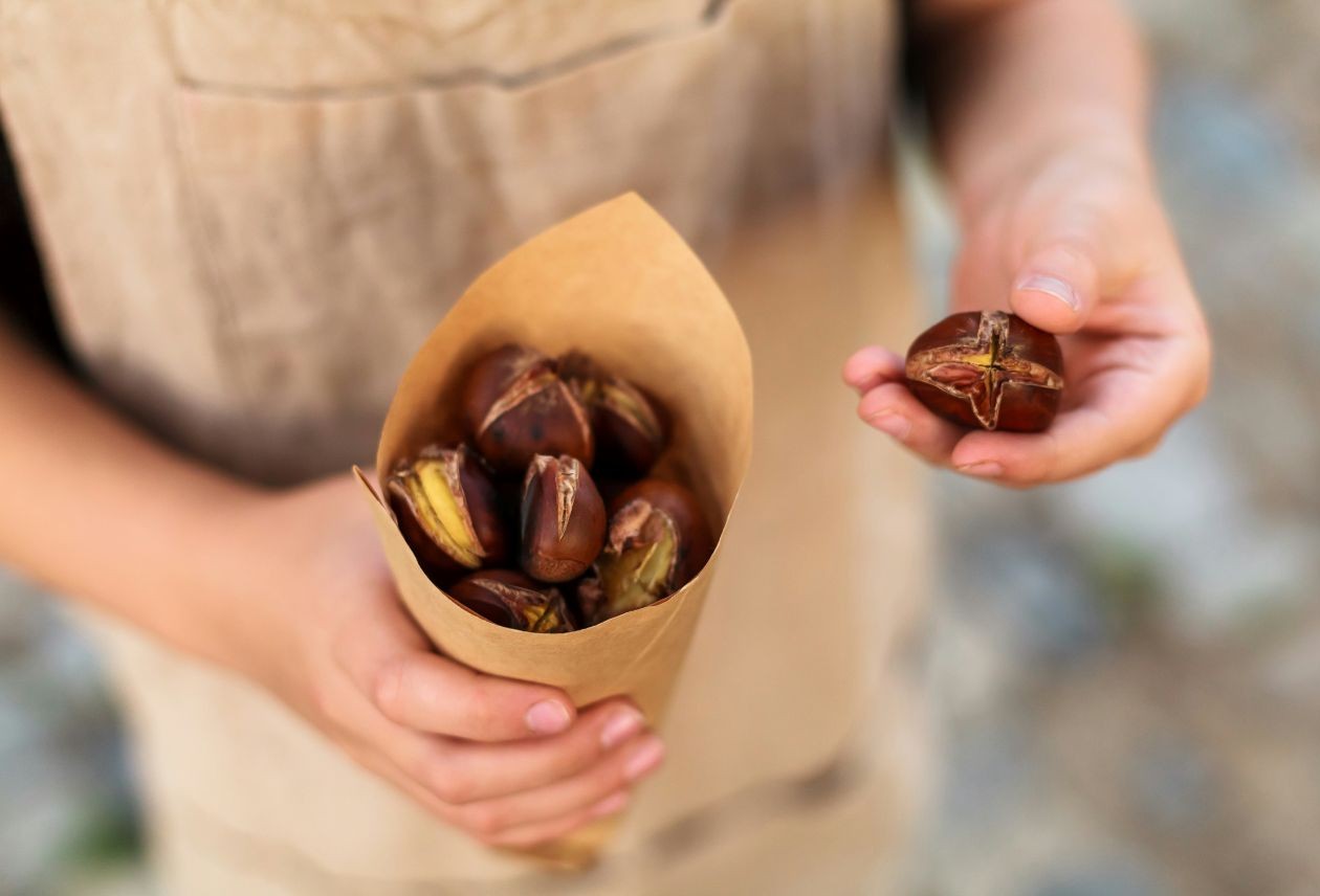Hands holding a paper cone filled with roasted chestnuts, while the other hand holds a single split, toasted chestnut.