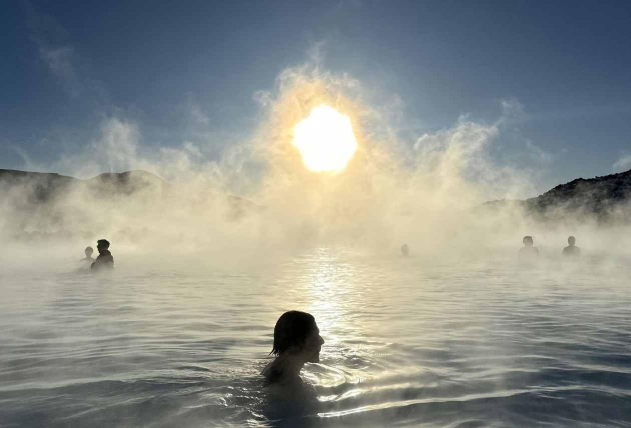 Silhouetted bathers in a steaming geothermal pool with the low sun breaking through mist and distant hills.