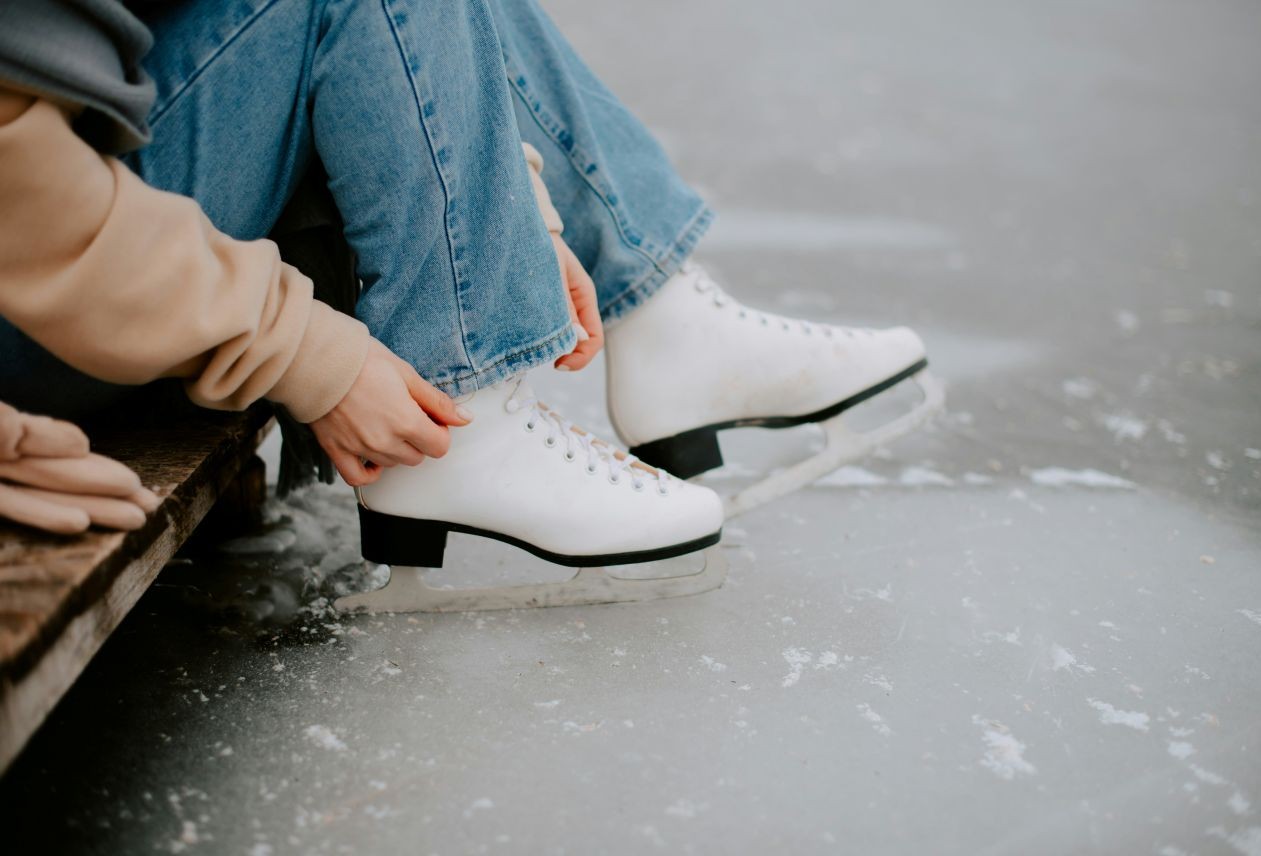Person in jeans and beige jacket sitting on a bench tying white figure skates over frozen ice.