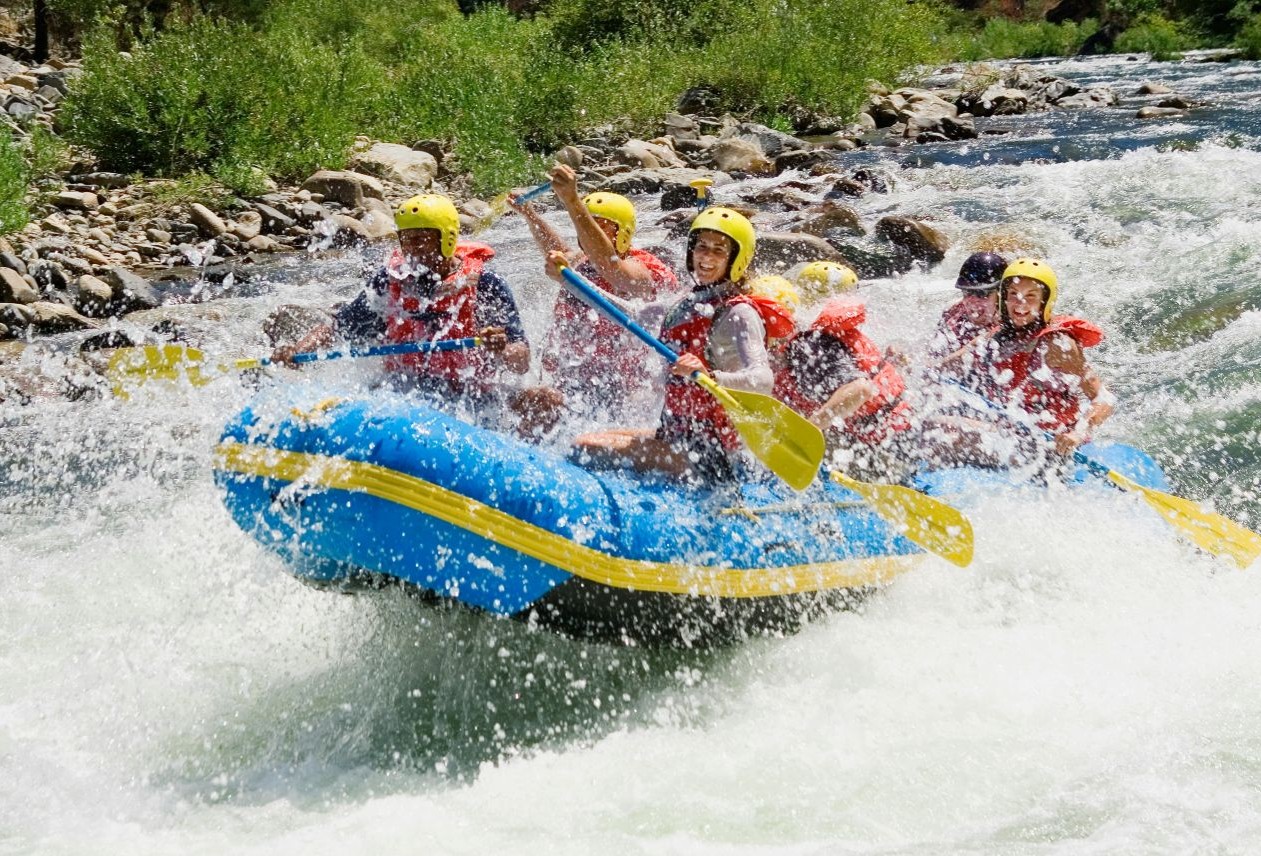 Group of rafters in yellow helmets and red life jackets paddling a blue inflatable raft through splashing whitewater rapids.