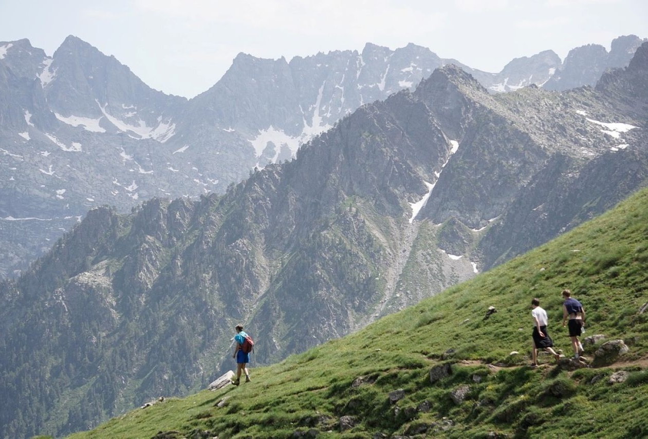 Three hikers walking on a grassy slope with a backdrop of jagged, snow-dusted mountain peaks under a cloudy sky.