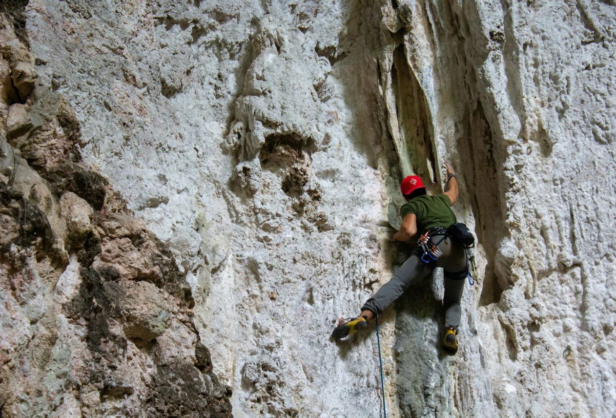 Rock climber in a red helmet and green shirt scaling a textured cliff face, using a crack for handholds and footholds.
