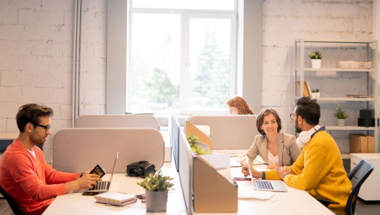 Open-plan office with four people working at desks. Three are engaged in conversation while one person works on a laptop. Plants and bright lighting.