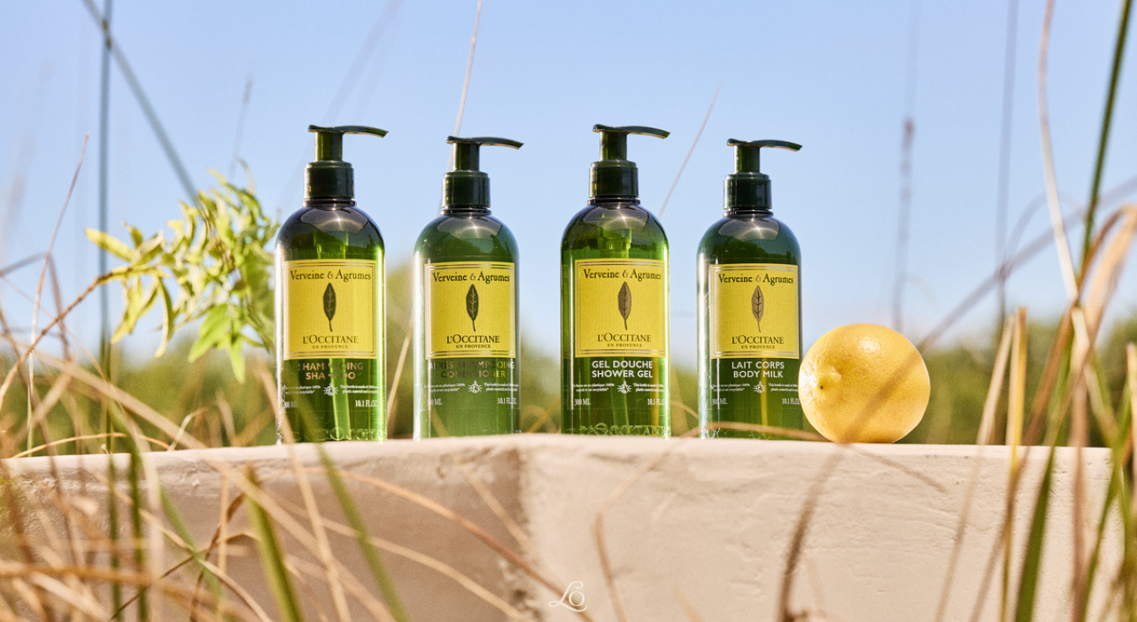 Four L'Occitane Verbena products on a stone ledge with a lemon and greenery, set against a clear blue sky background.