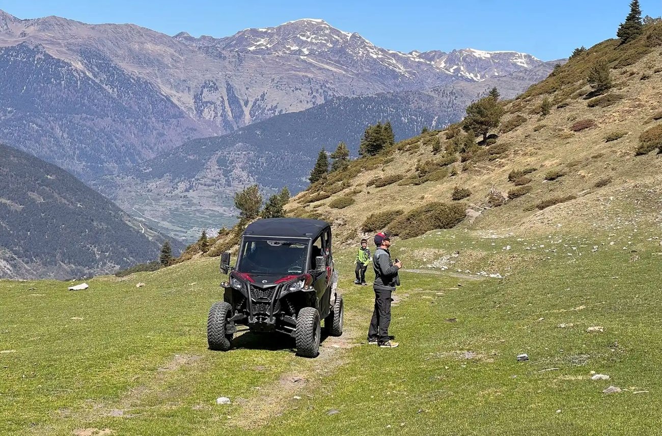 UTV parked on a grassy alpine slope with two people nearby and snow‑capped mountains in the background.