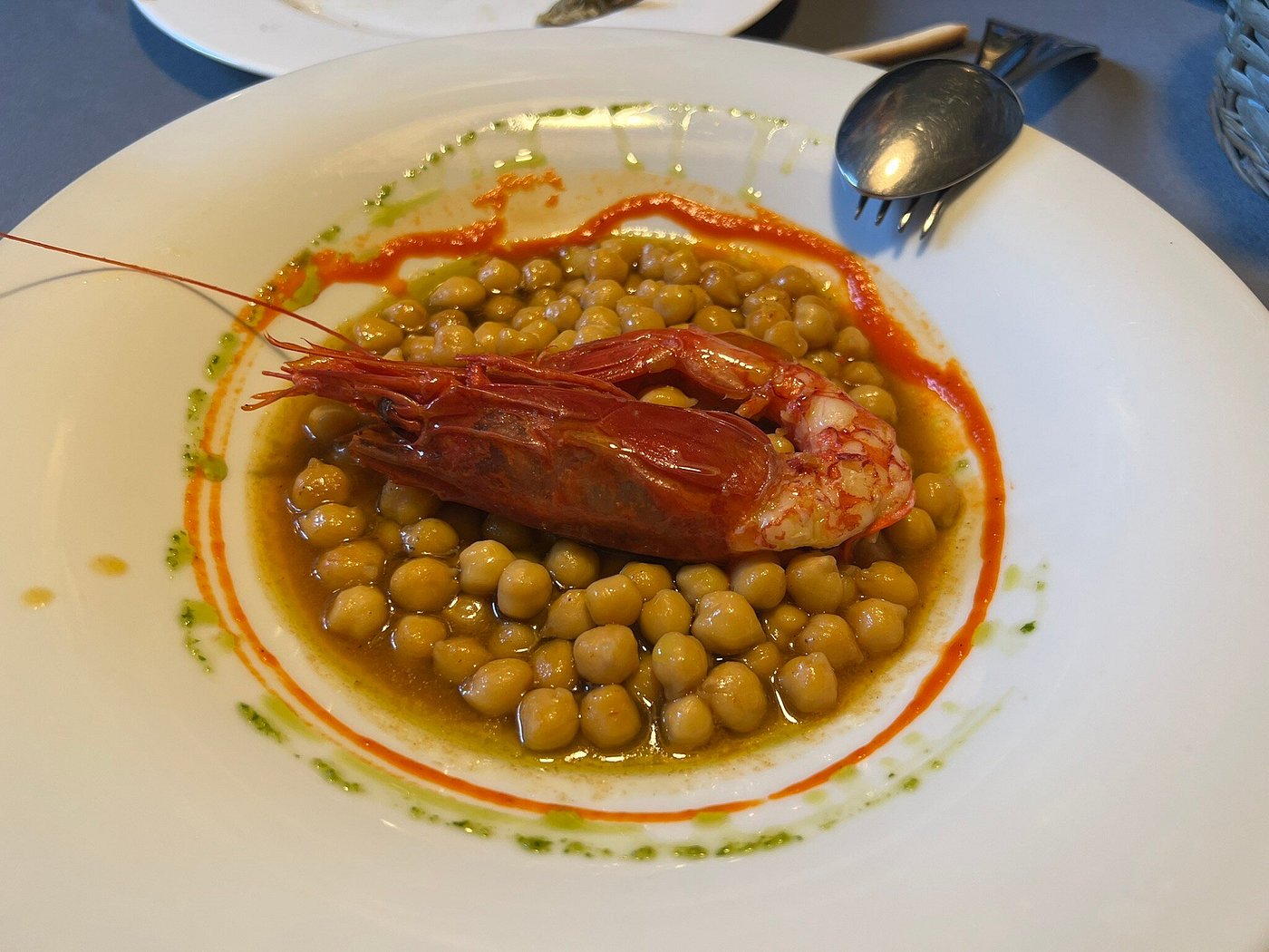 White bowl of chickpeas in broth topped with a whole cooked red prawn, drizzled with red and green sauce; spoon and fork at rim.