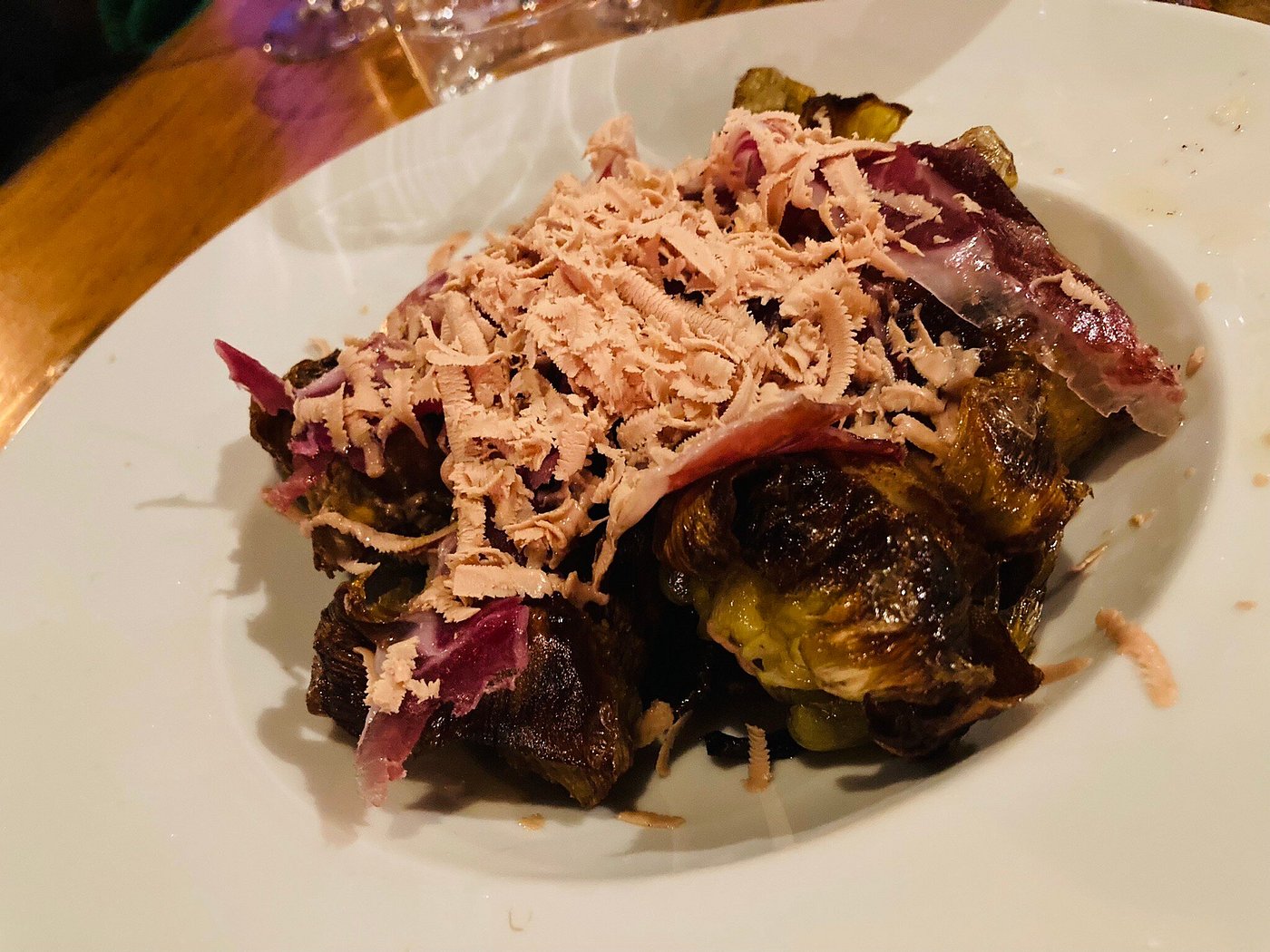 Roasted Brussels sprouts on a white plate topped with shredded pink cured meat and thin cheese shavings.