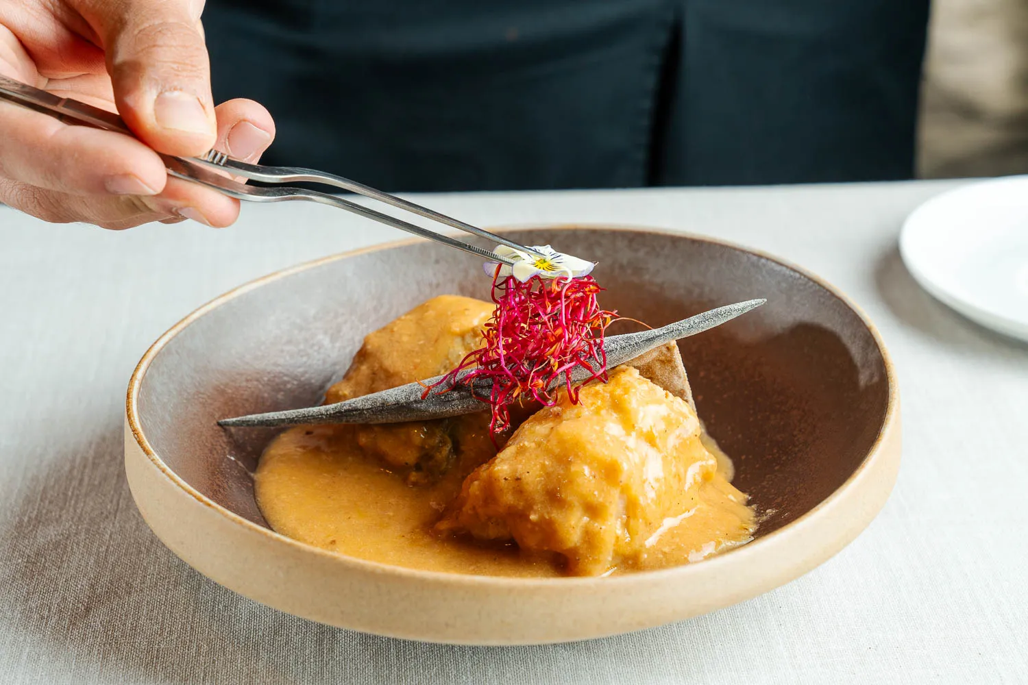 Chef using tweezers to place red microgreens on two sauce-covered dumplings in a brown bowl.