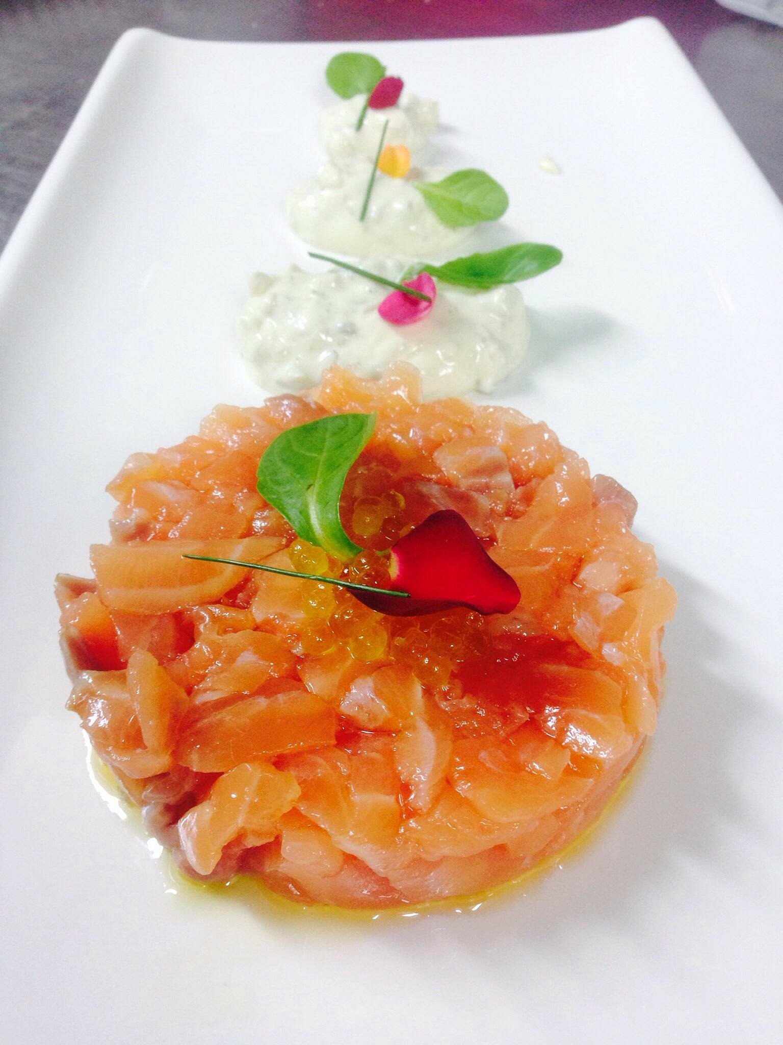 Round salmon tartare topped with microgreens and salmon roe, served with three dollops of herb cream on a long white rectangular plate.