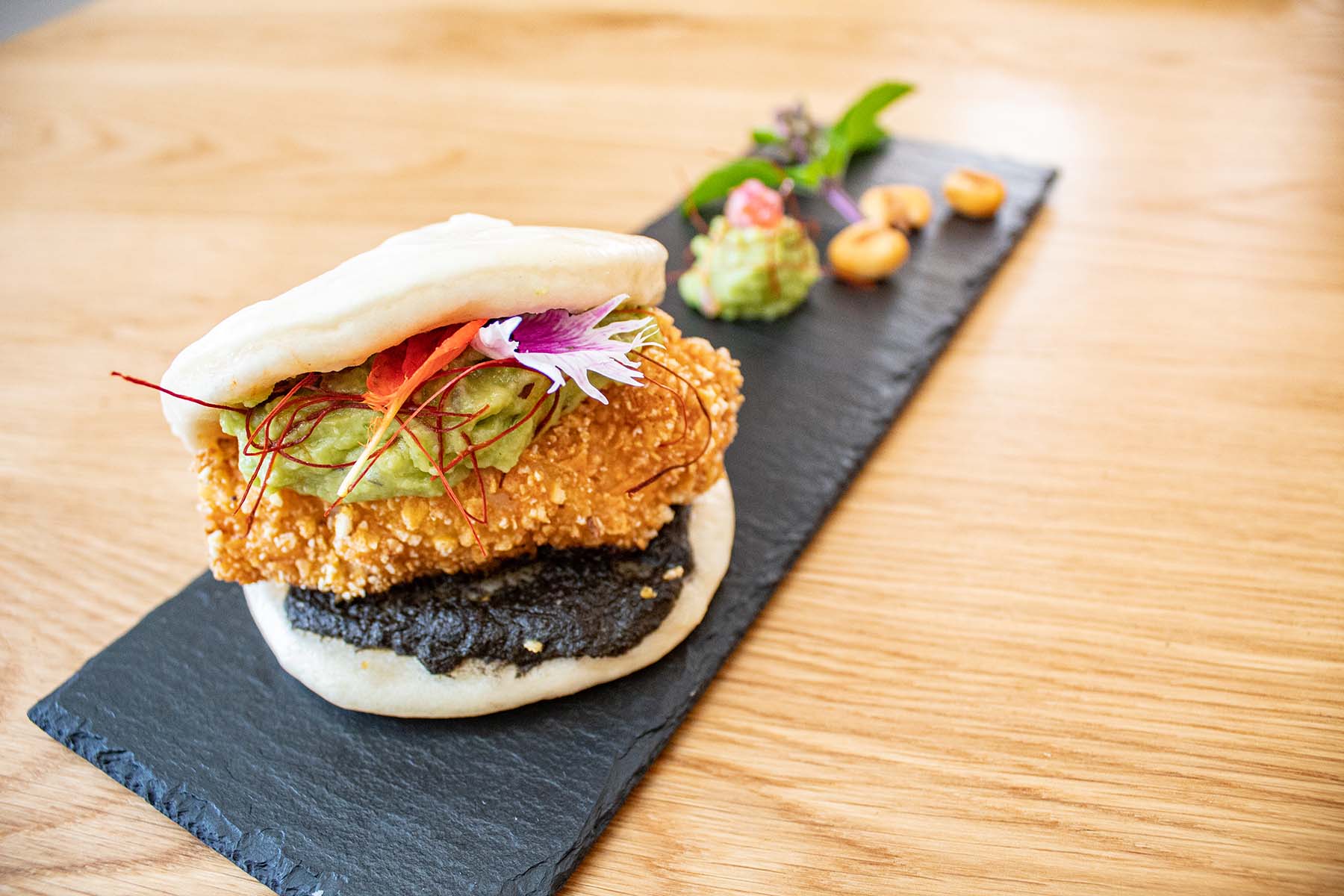 Steamed bao bun with crispy fried fillet, black sauce, avocado, microgreens and edible flower on a slate platter with small garnishes.