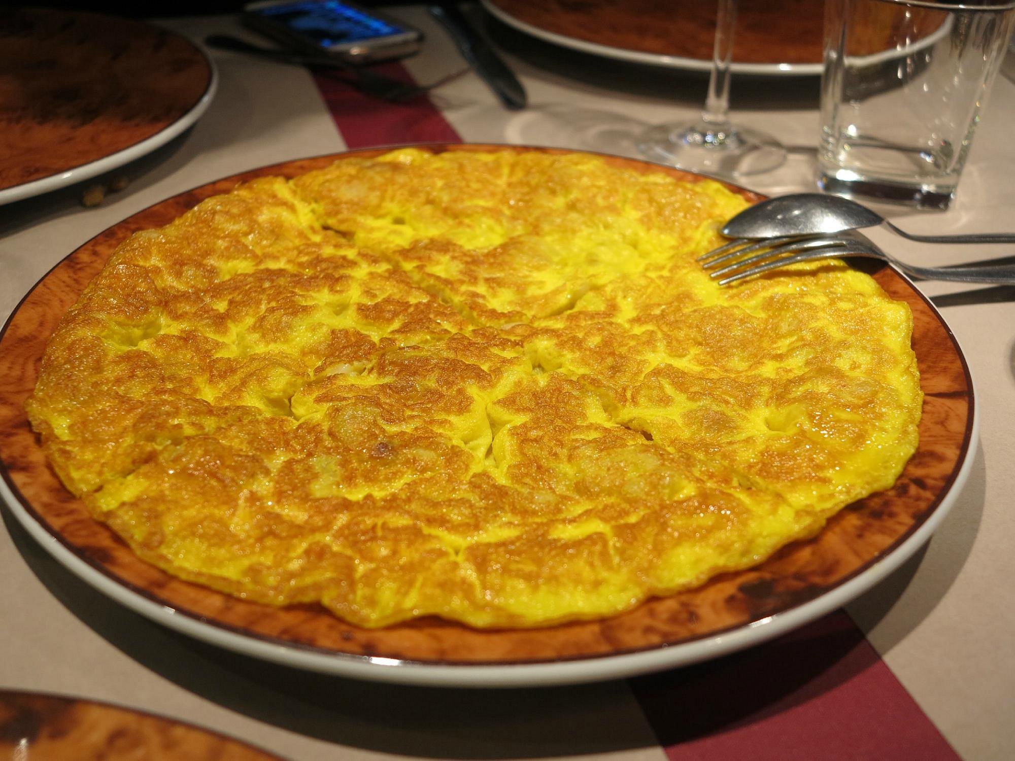 Large golden omelette on a brown-rimmed plate with a fork and spoon, set on a table with glasses nearby.