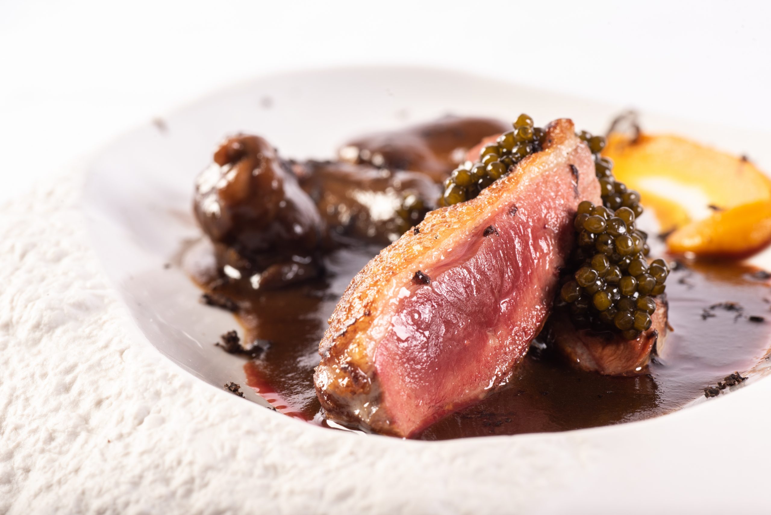 Medium-rare seared duck breast slices topped with black caviar, served on a glossy dark sauce with mushrooms and a roasted peach slice.