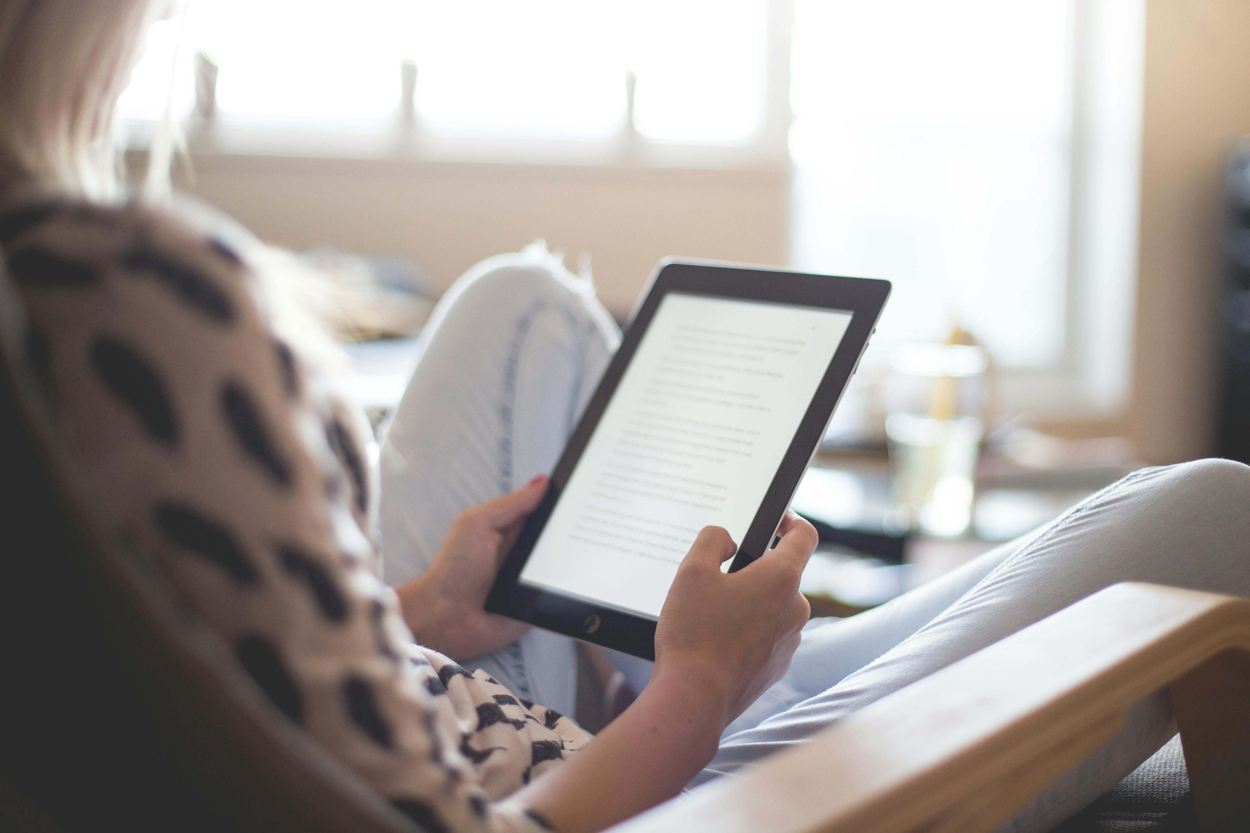 Person reading on a tablet while sitting in a chair, with a bright window in the background.