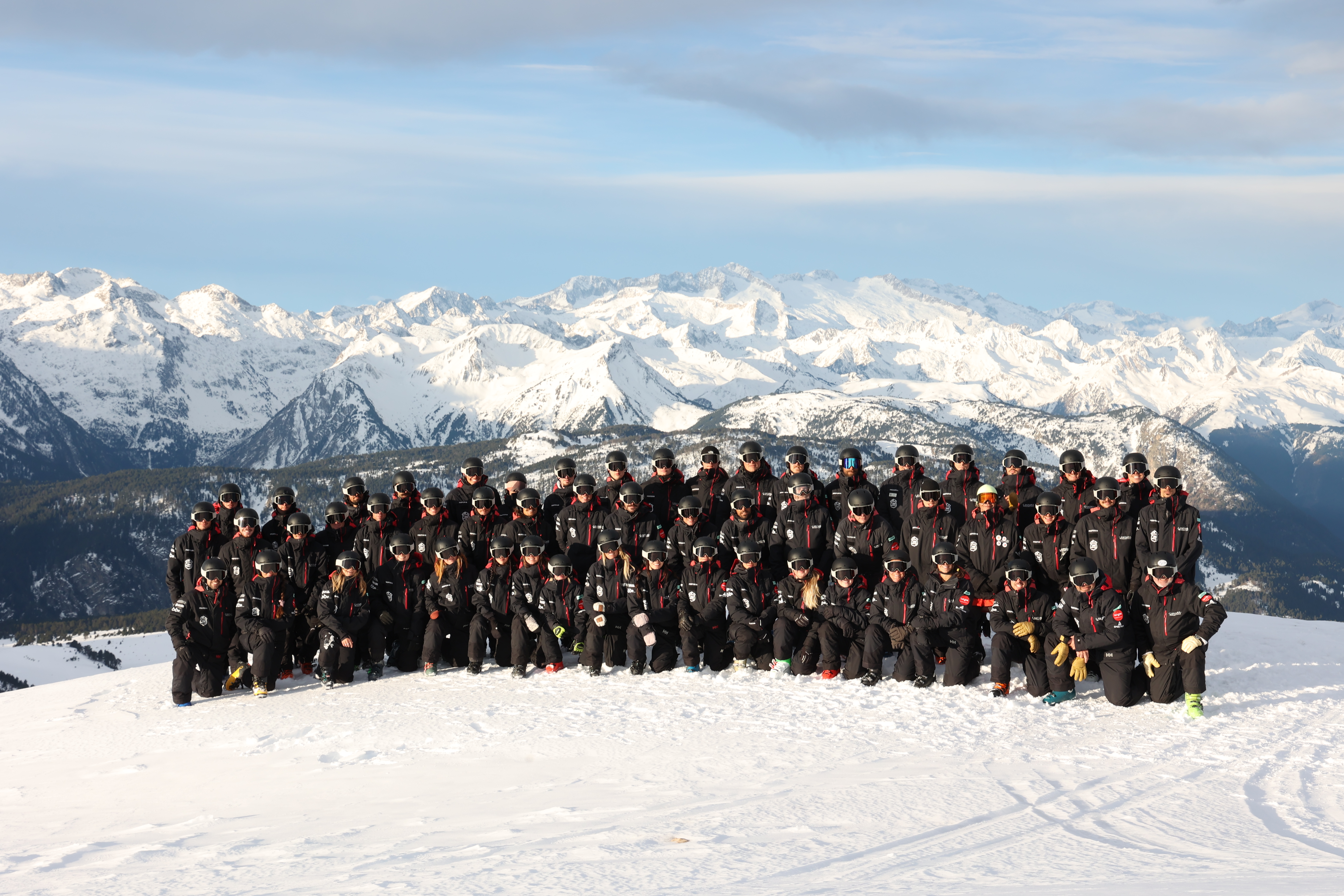 A large group of people in matching winter gear stand on a snowy mountain with snow-covered peaks in the background under a blue sky.