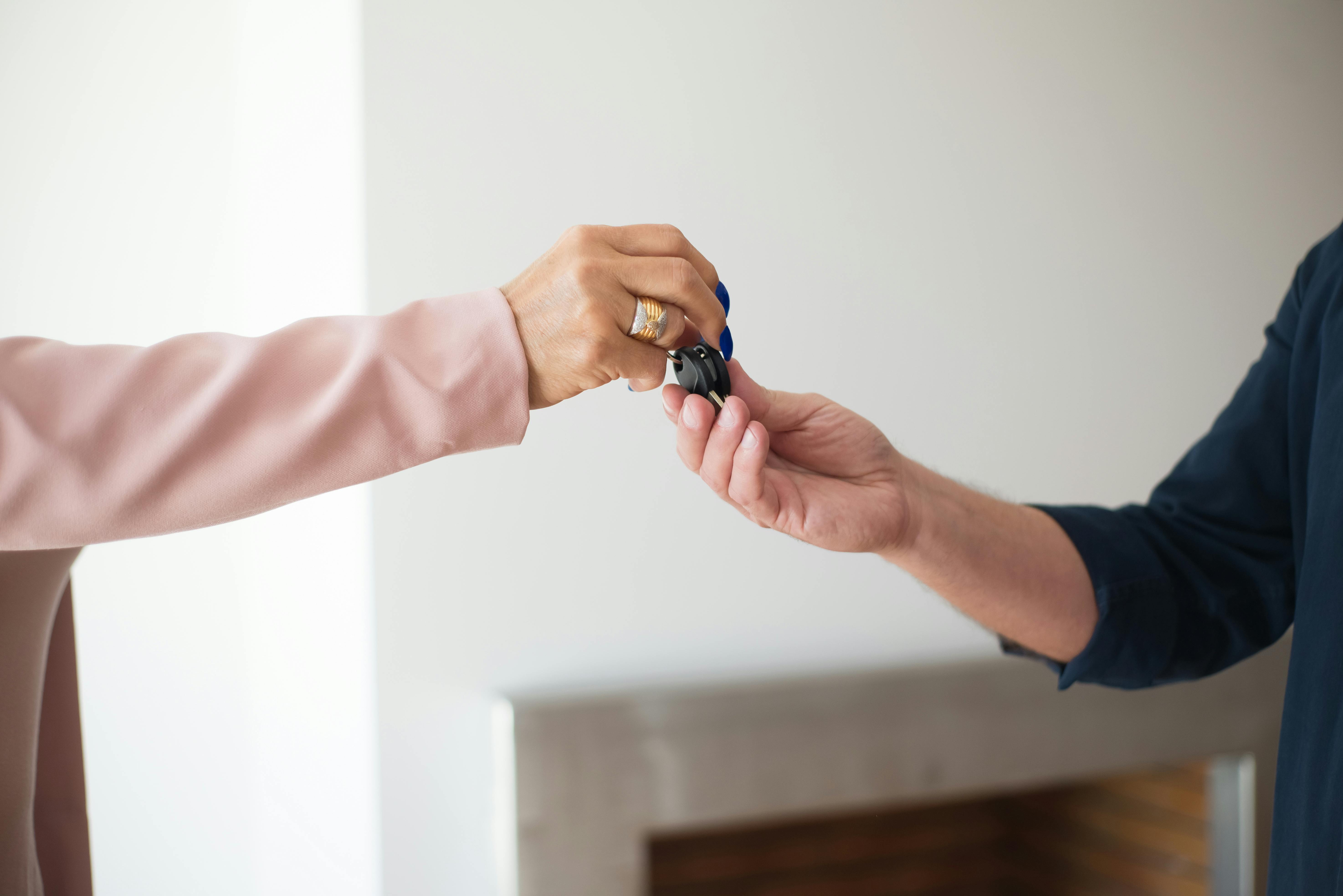 Two people exchanging car keys; one in a pink sleeve hands keys to another in a blue sleeve against a neutral background.