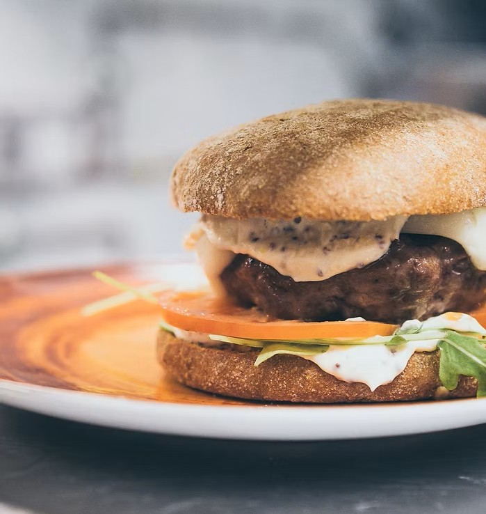 Close-up of a burger on a plate: toasted bun, beef patty, melting cheese, tomato, arugula and creamy sauce.