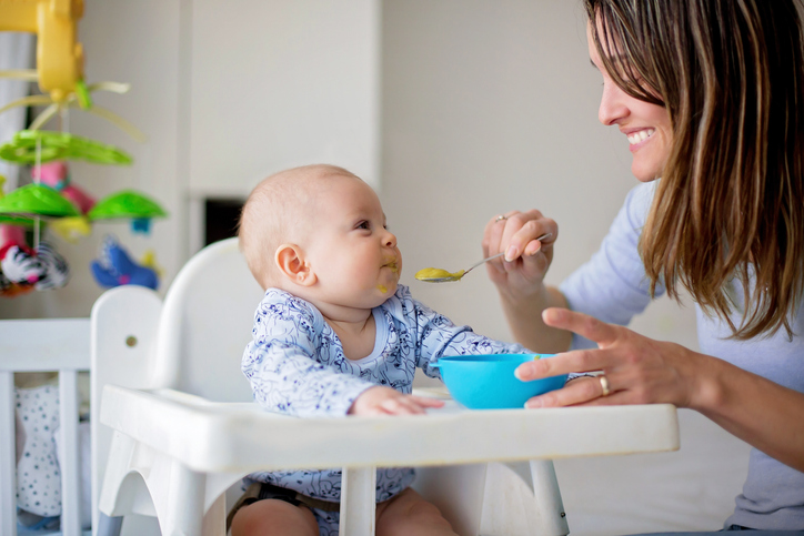 A smiling woman feeds a baby in a high chair. The baby is wearing a patterned outfit and looking eagerly at the spoon.