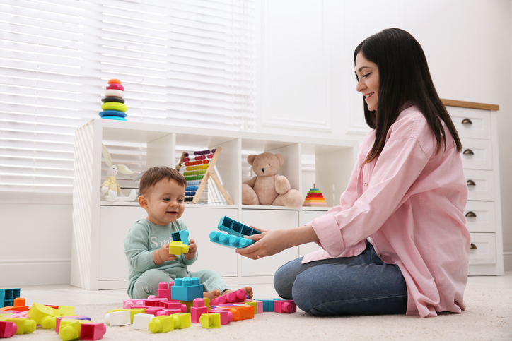 A woman and a baby sit on the floor playing with colorful toy blocks in a bright, cozy room with shelves and a teddy bear in the background.