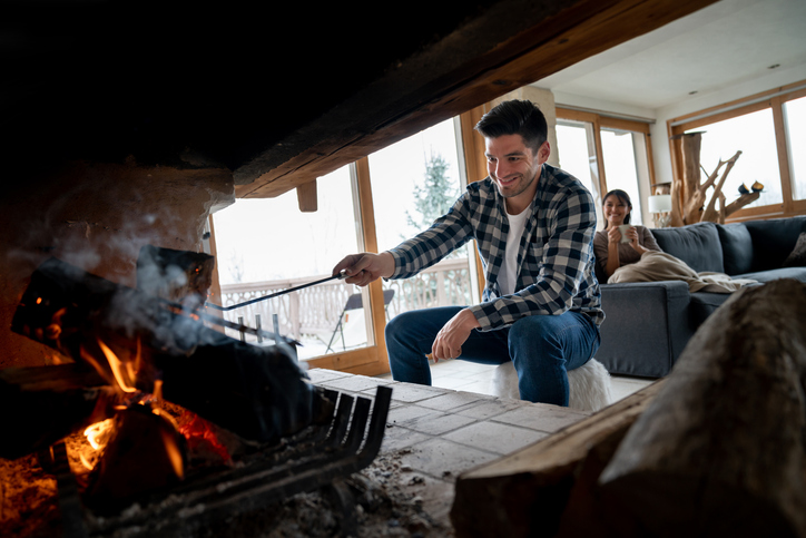 Man in plaid shirt tending a fireplace with a poker, while a woman sits on a couch in a cozy, light-filled room with large windows.