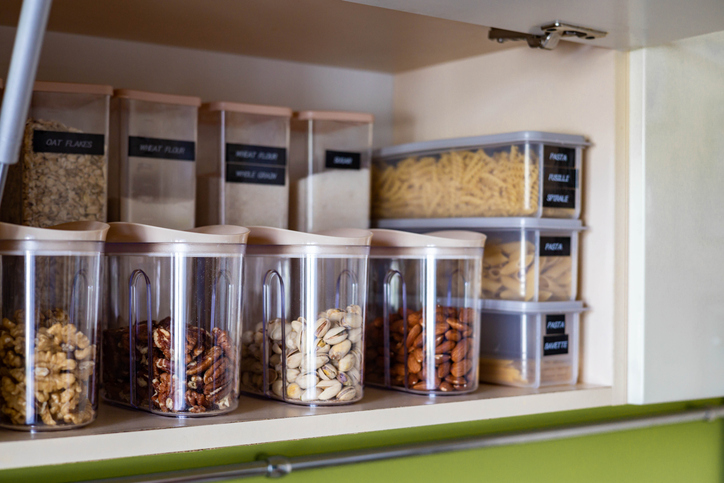 Open pantry shelf with labeled containers of assorted nuts, pasta, and grains, neatly organized in clear plastic bins.