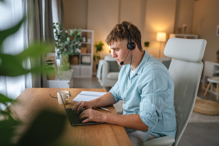 Person in a blue shirt sitting at a desk, wearing a headset, typing on a laptop. A notebook and a coffee mug are nearby.