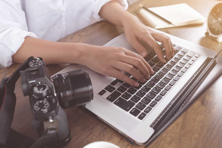 Hands typing on a laptop keyboard with a camera, clock, notebook, and cup on a wooden desk.