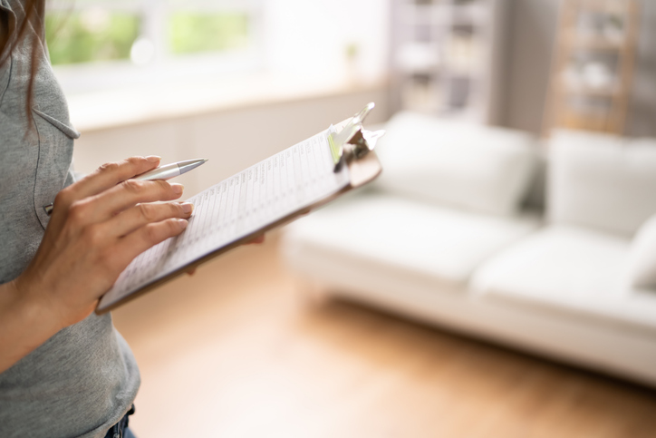 Person holding a pen and clipboard with paper, standing in a bright room with a white couch in the background.