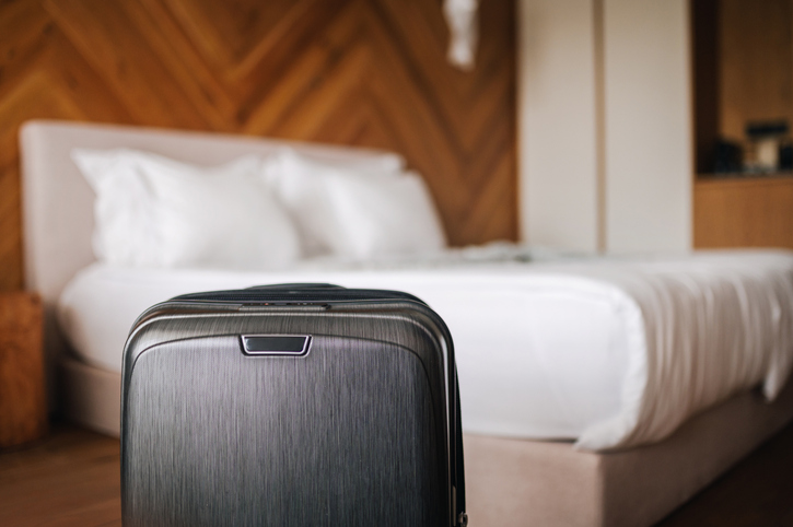 Suitcase in front of a neatly made bed with white linens in a cozy, wood-paneled hotel room.