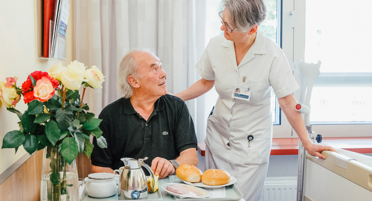 Eine freundliche Pflegekraft spricht mit einem Patienten, der am Tisch sitzt und eine ansprechende Mahlzeit vor sich hat.