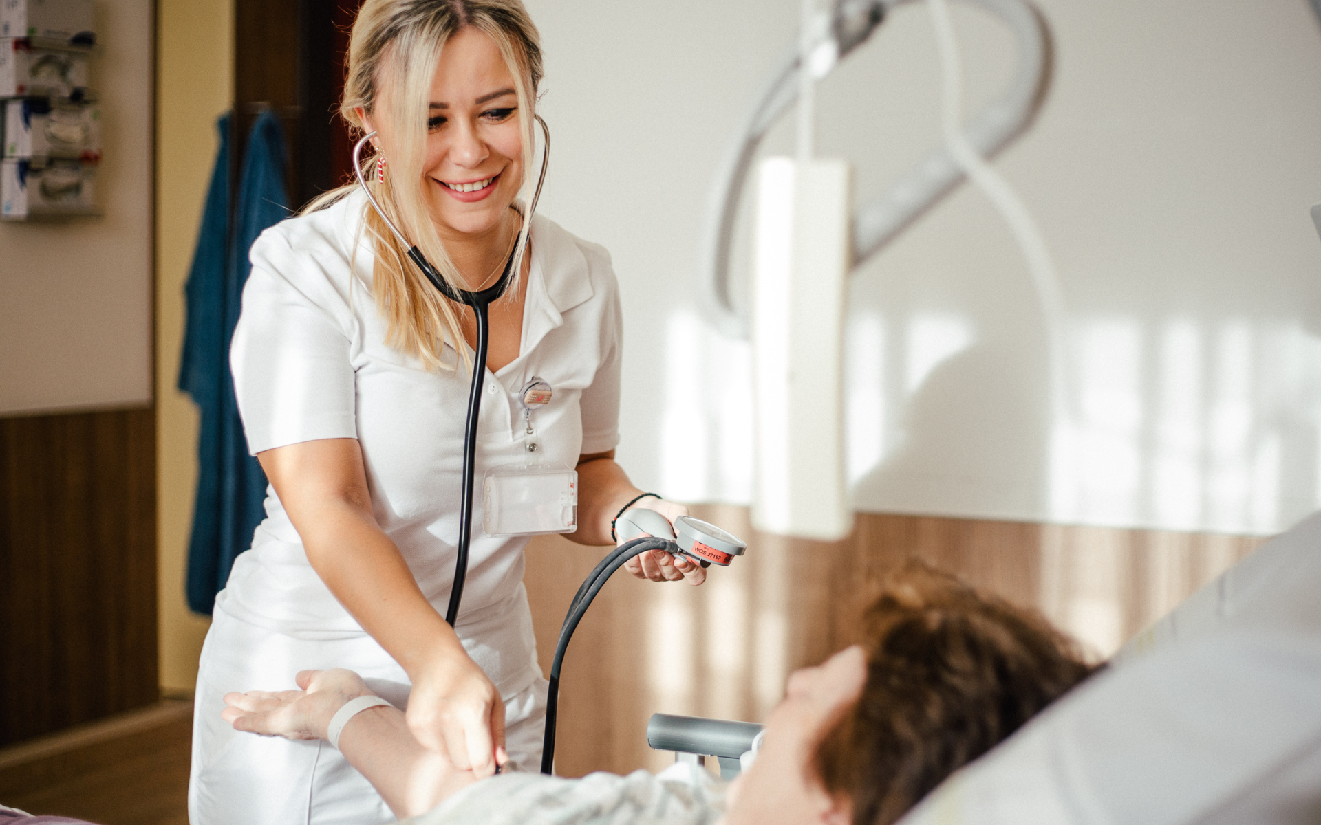 Eine Pflegekraft misst mit einem Blutdruckgerät den Blutdruck einer Patientin. Das Patientenzimmer ist hell und freundlich.