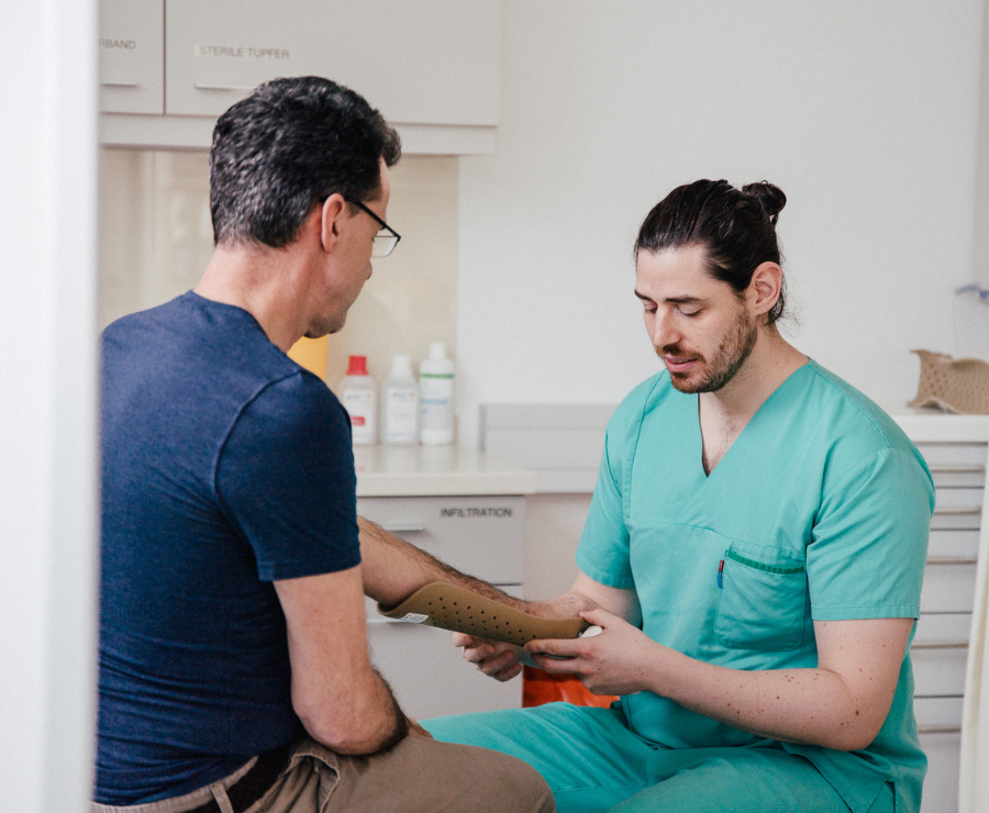 Ein freundlicher Arzt passt im Behandlungsraum der Akutambulanz einem Patienten eine Armschiene an.