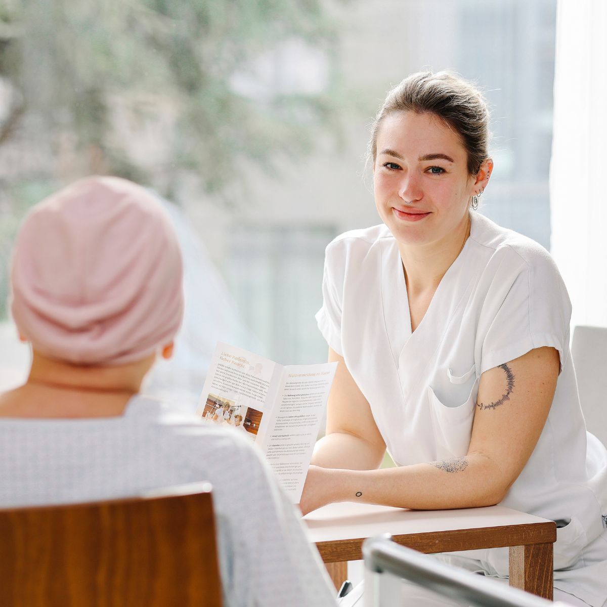 Eine Pflegeperson und eine Patientin der Onkologie mit Kopftuch sitzen sich gegenüber an einem Tisch.