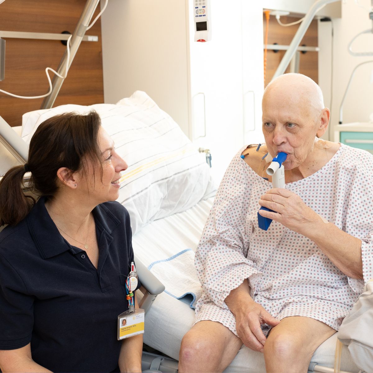 Eine Physiotherapeutin hockt vor dem Bett eines Patienten. Er sitzt im Bett und macht Atemübungen.